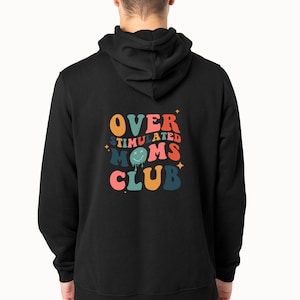 K&ouml;nnte beinhalten: Schwarzer Kapuzenpullover mit dem Text "OVERSTIMULATED MOMS CLUB" in einem Retro-Design. Das Design zeigt orange, rosa, blaue und gr&uuml;ne Schrift mit Sternakzenten. Der Hoodie hat eine Kapuze und lange &Auml;rmel.