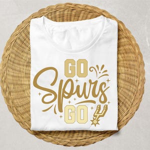 May include: A white t-shirt featuring gold graphic text that reads GO SPURS GO with decorative star accents and a small spur logo. The design is centered on the front of the shirt, which has a round neckline and short sleeves.
