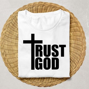 May include: White t-shirt with the words "TRUST GOD" in bold black letters and a black cross. The shirt is folded and placed on a round, woven, light brown surface.