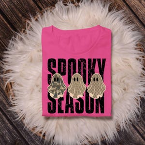 May include: A bright pink t-shirt featuring the black distressed text "SPOOKY SEASON" and three ghost illustrations. The ghosts have distinct patterns: one with a green camouflage print, one with a brown leopard print, and one with a tan checkerboard pattern.