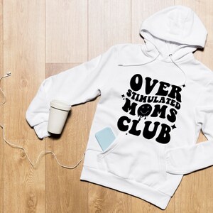 K&ouml;nnte beinhalten: Wei&szlig;er Kapuzenpullover mit dem Text "OVER STIMULATED MOMS CLUB" in Schwarz. Der Kapuzenpullover hat eine Vordertasche und eine Kapuze mit Kordelzug. Ein hellfarbiger, wiederverwendbarer Kaffeebecher und ein Telefon befinden sich in der Tasche. Wei&szlig;e kabelgebundene Kopfh&ouml;rer liegen neben dem Hoodie.