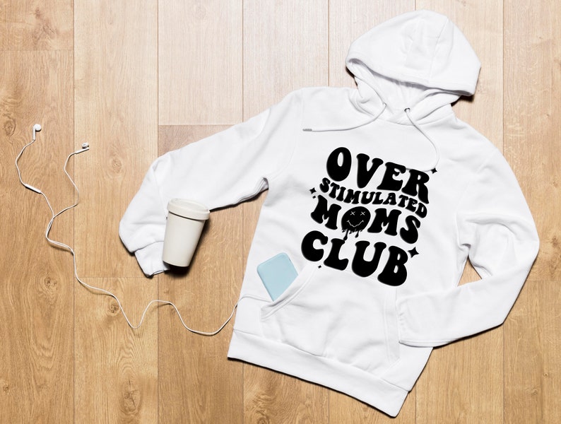 K&ouml;nnte beinhalten: Wei&szlig;er Kapuzenpullover mit dem Text "OVER STIMULATED MOMS CLUB" in Schwarz. Der Kapuzenpullover hat eine Vordertasche und eine Kapuze mit Kordelzug. Ein hellfarbiger, wiederverwendbarer Kaffeebecher und ein Telefon befinden sich in der Tasche. Wei&szlig;e kabelgebundene Kopfh&ouml;rer liegen neben dem Hoodie.