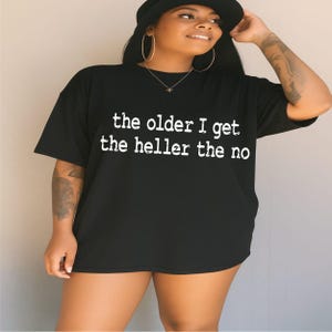 Può includere: Una t-shirt nera a maniche corte con una scritta bianca in stile usurato sul davanti che recita, the older I get the heller the no. Il capo presenta un girocollo e una vestibilità ampia e rilassata, offrendo un look casual e ben leggibile.