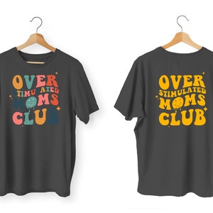 K&ouml;nnte beinhalten: Zwei anthrazitfarbene T-Shirts auf Holzb&uuml;geln. Die Vorderseite des ersten Shirts tr&auml;gt den Text "OVER STIMULATED MOMS CLUB" in Retro-Farben. Die R&uuml;ckseite des zweiten Shirts hat den gleichen Text in Gelb.