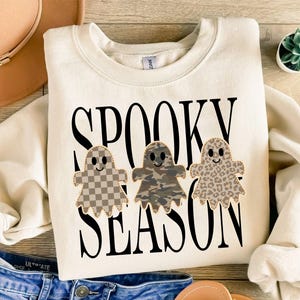 May include: A cream-coloured crewneck sweatshirt featuring the black text SPOOKY SEASON. Below the text are three ghost illustrations: one with a tan checkerboard pattern, one with a green and brown camouflage print, and one with a brown leopard print.