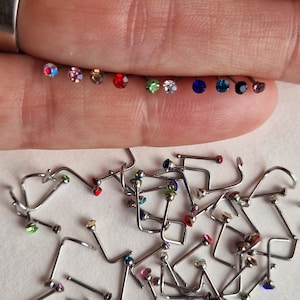 May include: A close-up of a selection of nose rings with various coloured gems. The gems are red, green, blue and pink. The nose rings are made of metal and have a curved shape.