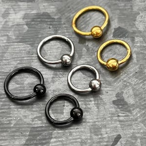May include: A selection of captive bead rings in gold, silver, and black. Each ring has a small, round bead to secure the closure. The jewellery is displayed on a textured, grey surface.