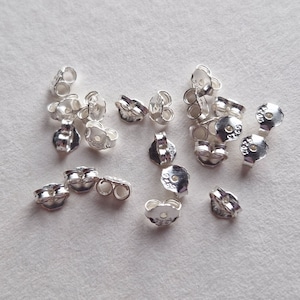 May include: A pile of silver earring backs. The backs are designed to fit over the post of an earring and have a small hole in the centre. The backs are marked with the number "925", which indicates that they are made of sterling silver.