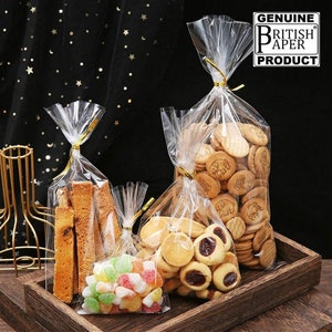 May include: Three clear plastic bags with gold twist ties filled with different treats. The bags are on a wooden surface. The largest bag is filled with biscuits, the middle bag is filled with breadsticks, and the smallest bag is filled with colourful sweets. The bags are labelled "GENUINE BRITISH PAPER PRODUCT".