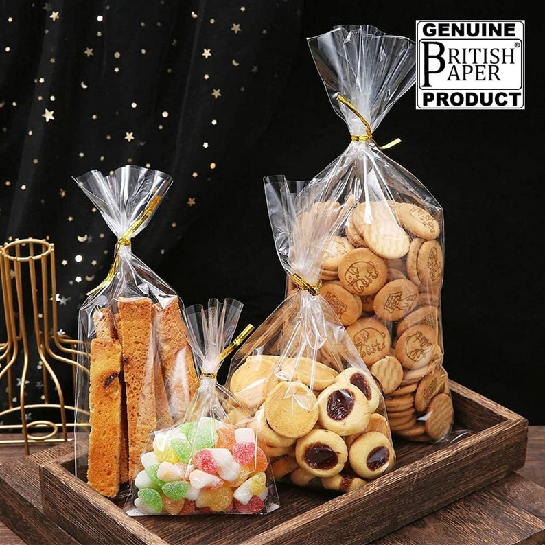 May include: Three clear plastic bags with gold twist ties filled with different treats. The bags are on a wooden surface. The largest bag is filled with biscuits, the middle bag is filled with breadsticks, and the smallest bag is filled with colourful sweets. The bags are labelled "GENUINE BRITISH PAPER PRODUCT".