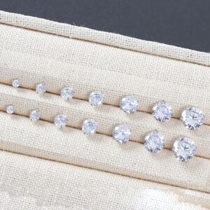May include: A selection of clear, round stud earrings in different sizes, presented on a beige fabric. The earrings are arranged in two rows, highlighting their sparkling facets. The largest earrings are approximately 0.8 cm in diameter.