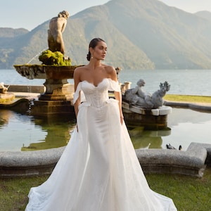 Peut inclure: Robe de mariée blanche à épaules dénudées avec une superposition de dentelle et une longue traîne. La robe présente un corsage ajusté et une jupe fluide. L'arrière-plan comprend une fontaine, un lac et des montagnes.
