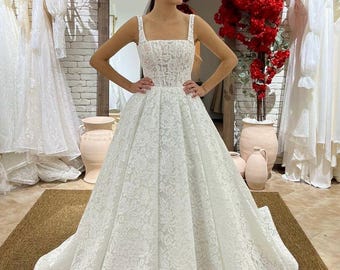May include: A white lace wedding dress with a square neckline and a full skirt. The dress is fitted at the bodice and flows out into a voluminous skirt.
