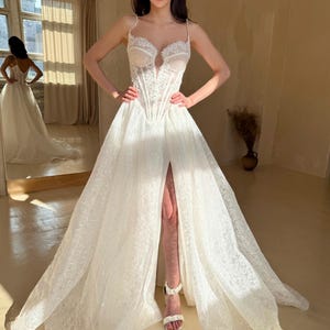 May include: A white lace wedding dress with a sweetheart neckline and a fitted bodice. The dress has a long, flowing skirt with a thigh-high slit and a train. The dress is decorated with delicate lace and beading.