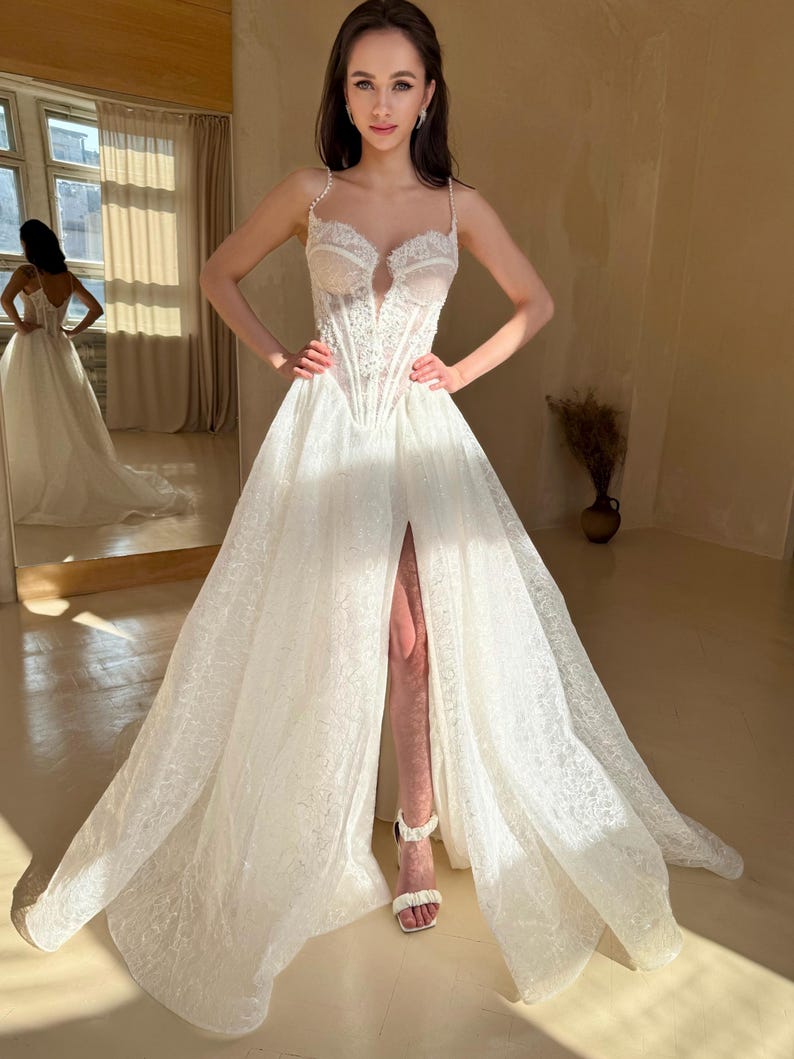 May include: A white lace wedding dress with a sweetheart neckline and a fitted bodice. The dress has a long, flowing skirt with a thigh-high slit and a train. The dress is decorated with delicate lace and beading.