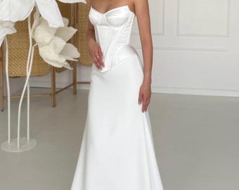 Puede incluir: Un vestido de novia blanco sin tirantes con un corpiño estructurado y una falda fluida. El vestido presenta un escote corazón y una cintura entallada. La modelo lleva un delicado collar. El fondo incluye grandes flores de papel blancas.