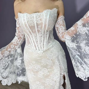 May include: White lace wedding dress with a corset bodice and off-the-shoulder sleeves. The dress features a high slit and intricate floral patterns throughout the fabric. The sleeves are long and flared, adding a dramatic flair to the bridal gown.