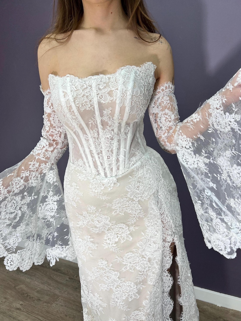 May include: White lace wedding dress with a corset bodice and off-the-shoulder sleeves. The dress features a high slit and intricate floral patterns throughout the fabric. The sleeves are long and flared, adding a dramatic flair to the bridal gown.