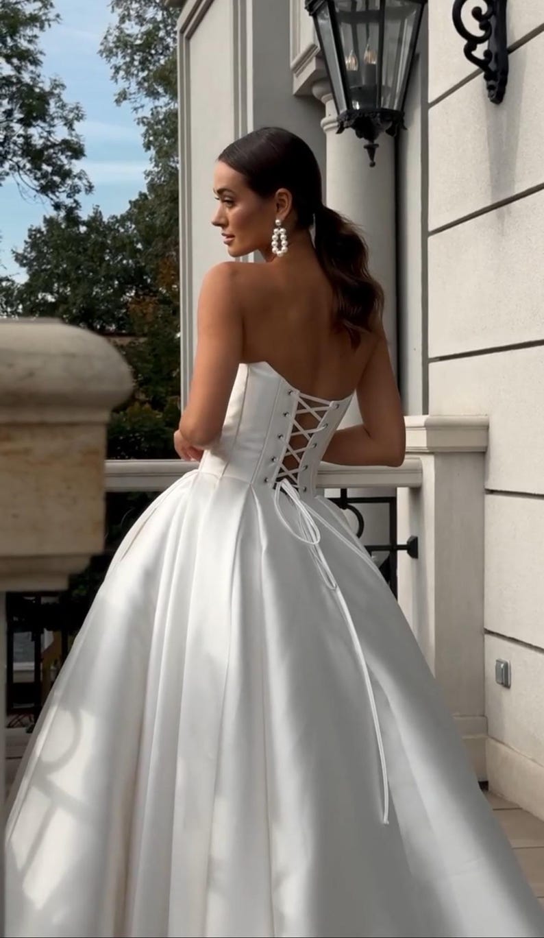 May include: A white strapless wedding dress with a lace-up back and a full skirt.