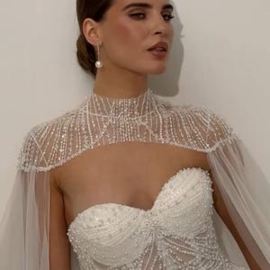 May include: A strapless white wedding dress with intricate beadwork and a high-neck cape adorned with beads. The dress features a sweetheart neckline and a flowing, sheer cape.