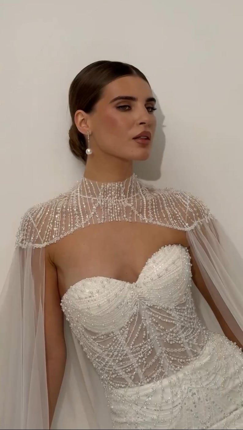 May include: A strapless white wedding dress with intricate beadwork and a high-neck cape adorned with beads. The dress features a sweetheart neckline and a flowing, sheer cape.