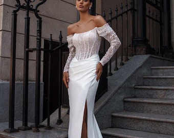 May include: A white off-the-shoulder wedding dress with a lace bodice and long sleeves. The skirt is a flowing, high-slit design. The dress is worn on a stone staircase next to a wrought iron fence.