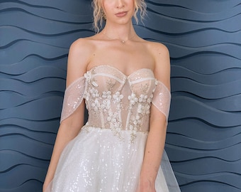 A-line wedding dress with cascading sleeves corset built-in cups 3D floral lace detailing, and a shimmering tulle skirt with floral pattern.