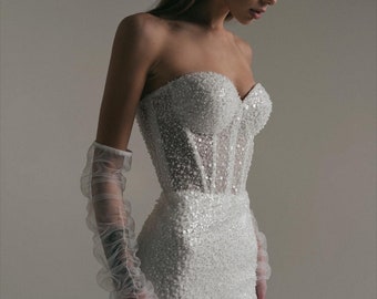 May include: A strapless white wedding dress with a fitted bodice and a flowing skirt. The dress is covered in sparkling sequins. Sheer, ruffled gloves extend up the arms. The overall look is elegant and glamorous.