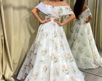 May include: A white off-the-shoulder gown with a floral pattern of pink and green roses. The dress has a full skirt and a fitted bodice. The model is standing in front of a mirror.