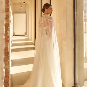 May include: A white wedding dress with a sheer cape, featuring a high neckline and intricate beadwork. The dress has a fitted bodice and a flowing skirt. The bride is standing in a sunlit hallway with brick and concrete walls.