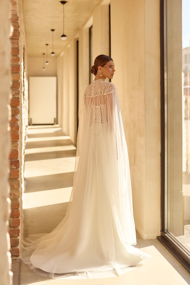 May include: A white wedding dress with a sheer cape, featuring a high neckline and intricate beadwork. The dress has a fitted bodice and a flowing skirt. The bride is standing in a sunlit hallway with brick and concrete walls.