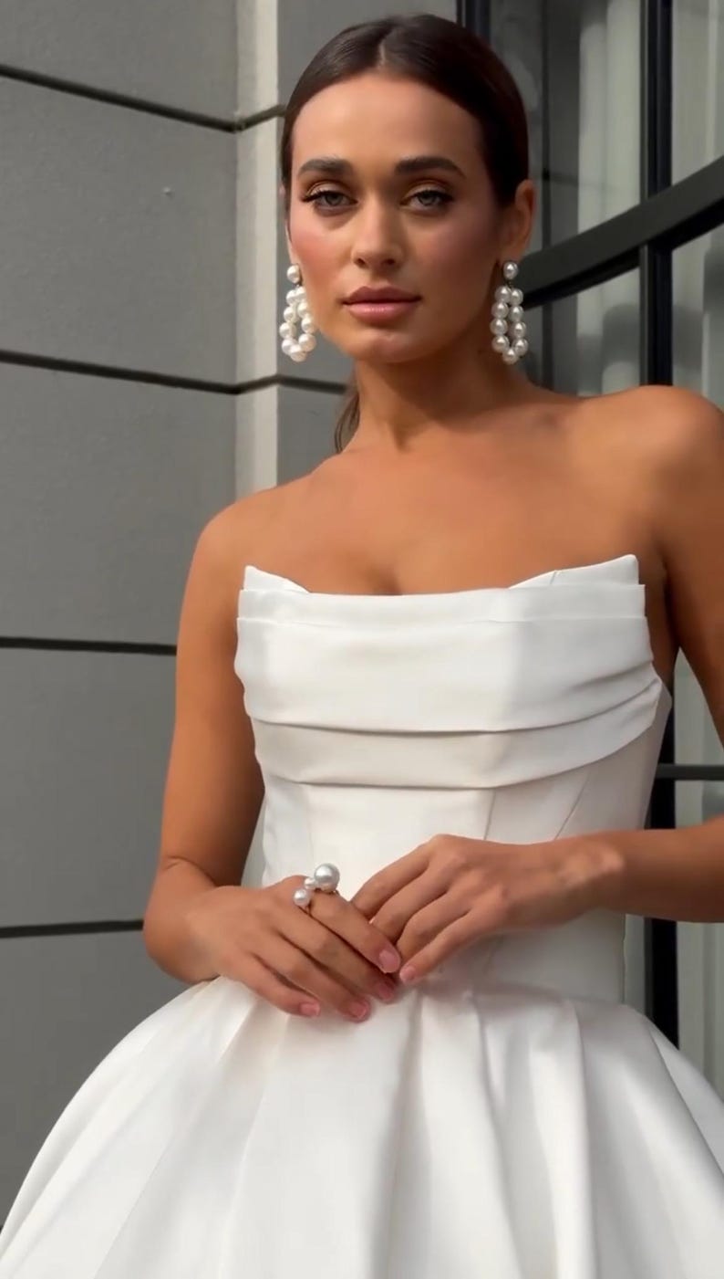 May include: A strapless white satin wedding dress with a flowing skirt. The dress has a simple, elegant design with a fitted bodice and a gathered skirt. The dress is accented with pearl jewelry.