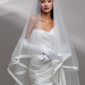 Peut inclure: Une mariée vêtue d'une robe de mariée en satin blanc, de longs gants et d'un voile transparent avec une bordure en satin. Le voile retombe sur les épaules et encadre le visage. Un collier et une bague délicats sont visibles.