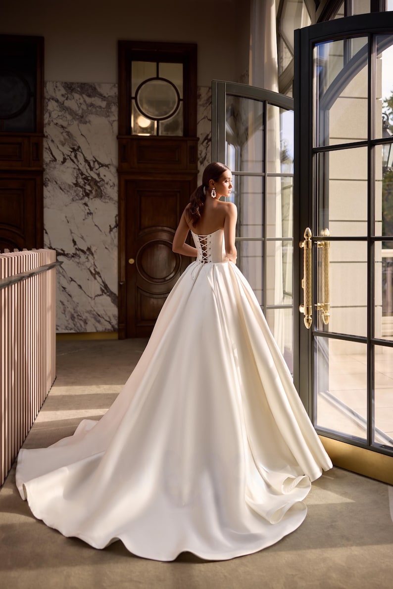 May include: A white satin wedding dress with a strapless bodice and a lace-up back. The dress has a full skirt and a long train.