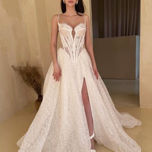 May include: A white lace wedding dress with a fitted bodice and a flowing skirt. The bodice has a sweetheart neckline and a corset-style design. The skirt has a high slit and a train. The dress is adorned with delicate lace details and sequins.