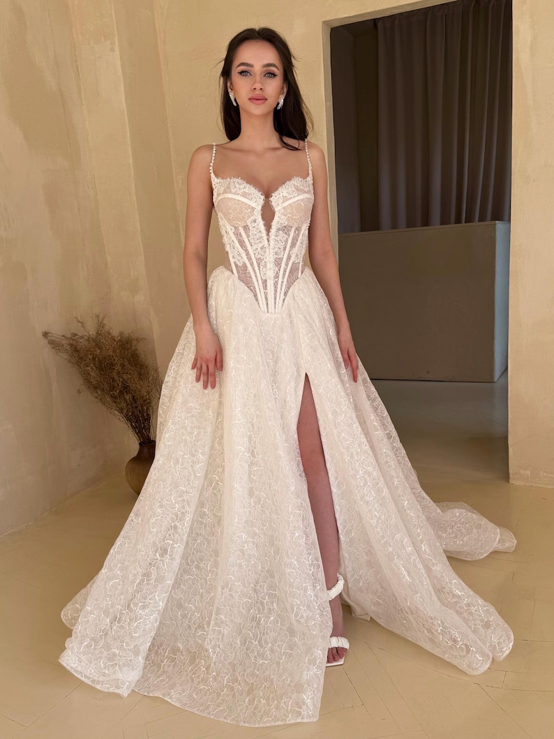 May include: A white lace wedding dress with a fitted bodice and a flowing skirt. The bodice has a sweetheart neckline and a corset-style design. The skirt has a high slit and a train. The dress is adorned with delicate lace details and sequins.