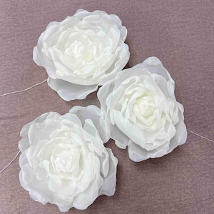 Op de afbeelding: Drie witte zijden pioenen met een draadsteel. De pioenen zijn in een rij op een bruin oppervlak gerangschikt.