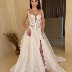 May include: A white lace wedding dress with a corset bodice and a flowing skirt. The skirt has a high slit on the left side and is adorned with delicate embroidery. The dress is designed for a formal occasion.