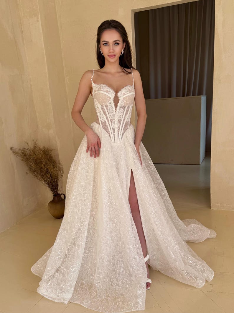 May include: A white lace wedding dress with a corset bodice and a flowing skirt. The skirt has a high slit on the left side and is adorned with delicate embroidery. The dress is designed for a formal occasion.
