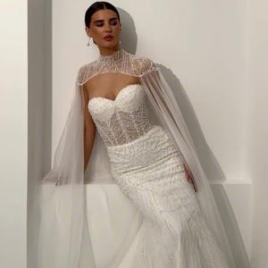 May include: A white, beaded wedding gown with a sweetheart neckline and a mermaid silhouette. The dress features intricate beadwork and a sheer, high-neck cape. The bride is leaning against a white wall.