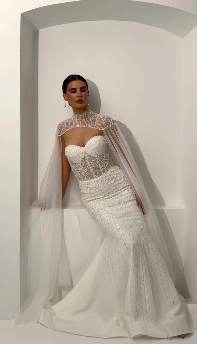 May include: A white, beaded wedding gown with a sweetheart neckline and a mermaid silhouette. The dress features intricate beadwork and a sheer, high-neck cape. The bride is leaning against a white wall.