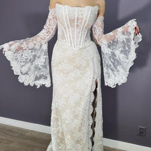 May include: A white lace wedding dress with an off-the-shoulder neckline and long, flared sleeves. The dress features a fitted bodice with vertical boning and a high slit on the side of the skirt. The dress has a long train.