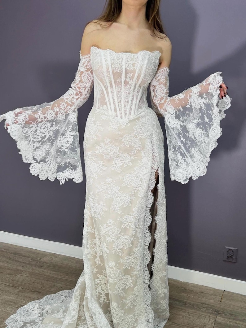 May include: A white lace wedding dress with an off-the-shoulder neckline and long, flared sleeves. The dress features a fitted bodice with vertical boning and a high slit on the side of the skirt. The dress has a long train.