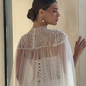 May include: A white wedding dress with a beaded cape. The cape is adorned with intricate beadwork and drapes over the shoulders. The dress features a corset back and a flowing skirt, creating an elegant silhouette. The overall design is classic and romantic.