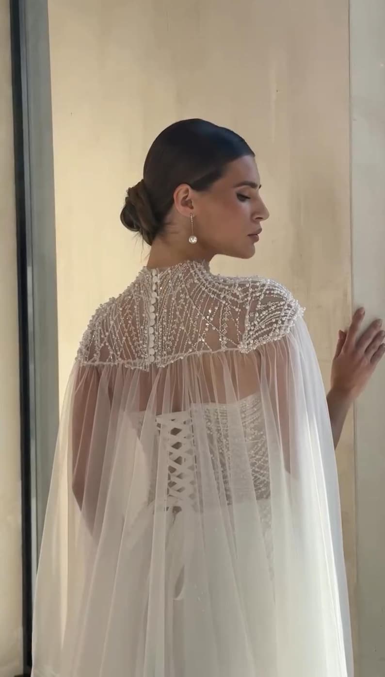 May include: A white wedding dress with a beaded cape. The cape is adorned with intricate beadwork and drapes over the shoulders. The dress features a corset back and a flowing skirt, creating an elegant silhouette. The overall design is classic and romantic.