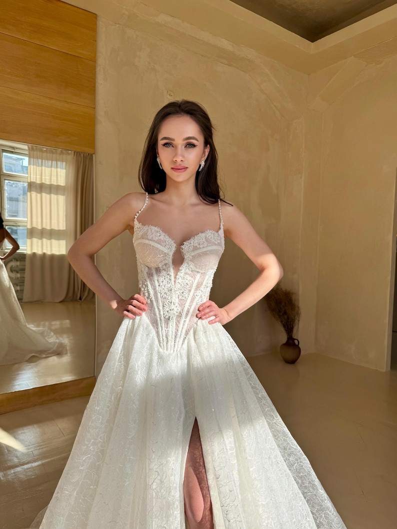 May include: A white wedding dress with a lace bodice and a flowing skirt. The skirt has a slit on the left side. The dress is embellished with sequins and beads.
