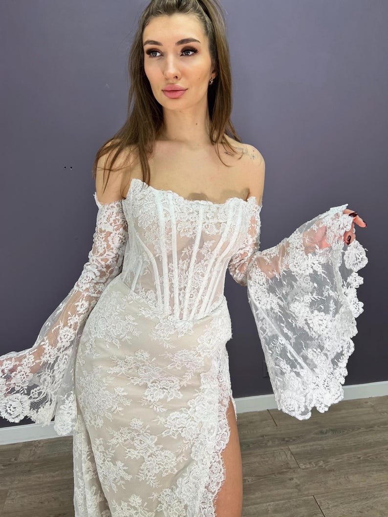 May include: White lace off-the-shoulder wedding dress with long, flared sleeves. The dress features a fitted bodice and a skirt with a high slit. The intricate lace detailing covers the entire dress, creating a romantic and elegant look.