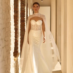 May include: A white, beaded wedding gown with a sweetheart neckline and a high-neck cape. The gown has a fitted bodice and a flowing skirt. The cape is sheer and drapes over the shoulders. The dress is worn in a sunlit hallway.