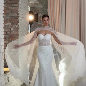May include: A white wedding gown with a fitted bodice and a flowing mermaid skirt. The dress is embellished with sparkling details. A sheer, beaded cape drapes over the shoulders, adding a touch of elegance. The model is standing in a studio setting.