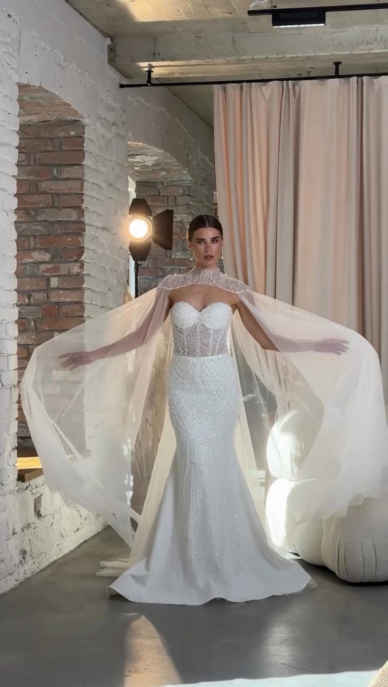 May include: A white wedding gown with a fitted bodice and a flowing mermaid skirt. The dress is embellished with sparkling details. A sheer, beaded cape drapes over the shoulders, adding a touch of elegance. The model is standing in a studio setting.
